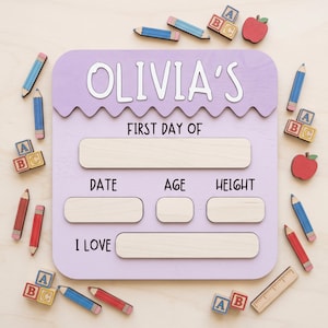 May include: A light purple wooden sign with the text OLIVIA'S FIRST DAY OF, featuring blank spaces for date, age, height, and interests. The sign is surrounded by small wooden cutouts of blue and red pencils, alphabet blocks, and red apples.
