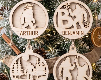 May include: Four circular light-colored wooden Christmas ornaments with cut-out designs. The top left features a Bigfoot in a Santa hat labeled Arthur. The top right shows a Bigfoot with the letter B labeled Benjamin. The bottom left depicts a forest scene with a Bigfoot labeled Cedric. The bottom right shows two Bigfoots, one making a peace sign, labeled Daisy.