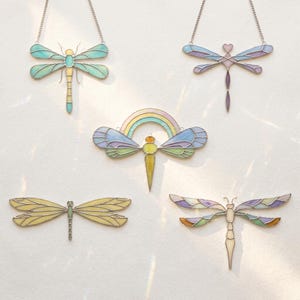 May include: Five stained glass dragonfly pendants in various pastel colors, including blue, yellow, and purple. Each dragonfly has a silver chain for hanging. The designs include a rainbow and heart accents.