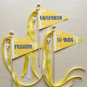 Könnte beinhalten: Drei gelbe dreieckige Wimpel an Holzstäben, jeweils mit weißen Blumenmustern und langen Bändern. Die Flaggen sind mit schwarzem Text beschriftet, der Preschool, Kindergarten und 1st Grade lautet, akzentuiert durch kleine weiße gänseblümchenförmige Perlen.