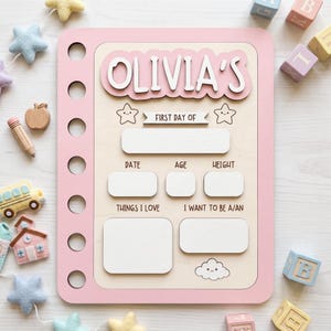 May include: A rectangular wooden sign with a pink border and a light wood centre, featuring the text OLIVIA'S, FIRST DAY OF, DATE, AGE, HEIGHT, THINGS I LOVE, and I WANT TO BE A/AN, with blank white spaces for writing information and star and cloud icons.