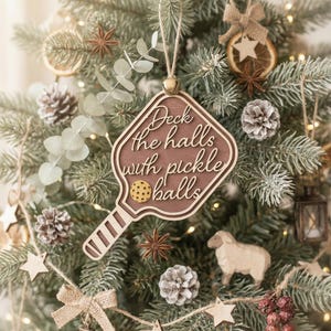 May include: Wooden Christmas ornament shaped like a pickleball paddle, with the text "Deck the halls with pickle balls." The ornament is brown with a light-colored border and a small gold bell. The ornament hangs on a Christmas tree decorated with pinecones and star-shaped ornaments.