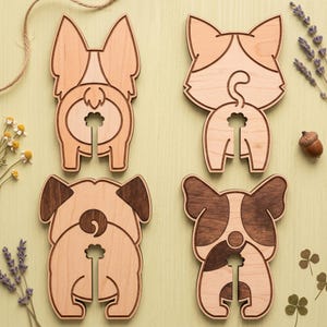 Puede incluir: Cuatro posavasos de madera con traseros de perros de varias razas. Cada posavasos es de color marrón claro con detalles en marrón oscuro, con un diseño único. Los posavasos están dispuestos sobre una superficie verde claro.