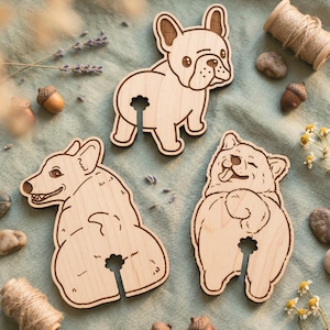 Op de afbeelding: Drie houten onderzetters in de vorm van honden: een Franse bulldog, een Corgi en een beerachtige hond. Elk heeft een sleuf voor een wijnglas. De onderzetters zijn lichtbruin met geëtste details, op een lichtgroene stof.