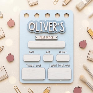 May include: A light blue wooden sign with five circular holes at the top. The text reads OLIVER'S, FIRST DAY OF, DATE, AGE, HEIGHT, THINGS I LOVE, and I WANT TO BE A/AN. It features blank white rectangular spaces for writing information.