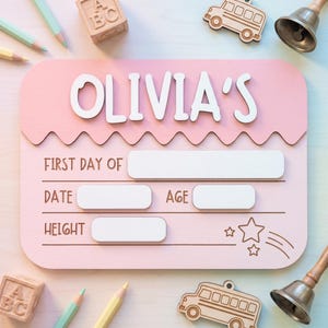 May include: A rectangular pink wooden sign with rounded corners featuring the white text OLIVIA'S at the top. Below, the sign includes engraved labels for FIRST DAY OF, DATE, AGE, and HEIGHT, with blank white rectangular spaces for writing.