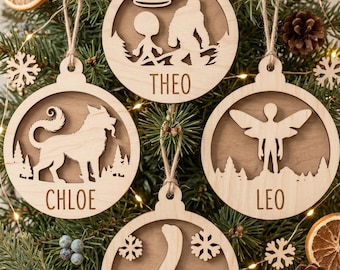 May include: Four circular light-colored wooden ornaments with cut-out designs. One features a UFO, alien, and Bigfoot with the name THEO. One shows a dog with the name CHLOE. One depicts a winged figure with the name LEO. One displays a long-necked dinosaur with the name MAYA.