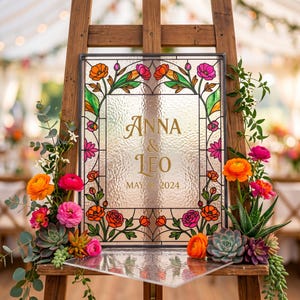 May include: A stained glass wedding sign with the names "Anna & Leo" and the date "May 18, 2024" in gold lettering. The sign is framed with colorful floral designs and rests on a wooden easel, decorated with flowers and succulents.