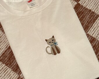 May include: White t-shirt with a brown embroidered cat wearing a blue bow.