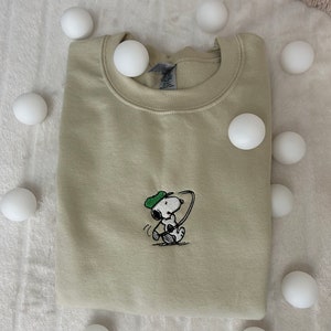 Op de afbeelding: Een lichtgroen sweatshirt met een cartoon afbeelding van Snoopy die golf speelt. Het sweatshirt is gevouwen en omringd door witte ballen.