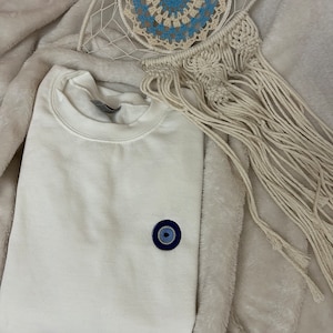May include: White sweatshirt with a blue and black evil eye charm on the front. The sweatshirt is folded and lying on a white fuzzy blanket.