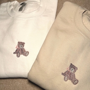 Puede incluir: Dos sudaderas, una blanca y otra beige, con un oso de peluche marrón bordado con un lazo rosa en cada una.