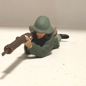 May include: A miniature figurine of a soldier in a prone position, aiming a rifle. The soldier wears a green helmet and uniform, with a brown rifle. The figure is painted with realistic details, suggesting a historical or military theme.