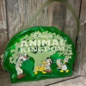 Op de afbeelding: Een groene plastic tas met een doorzichtige band. De tas heeft een afbeelding van Mickey Mouse, Goofy en Donald Duck in safari-outfits. De tas heeft de tekst "Disney's Animal Kingdom" in witte letters.