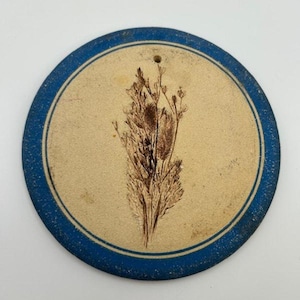 Puede incluir: Un posavasos redondo de cerámica de color crema con un borde azul. Un diseño de flor prensada y seca se encuentra en el centro.