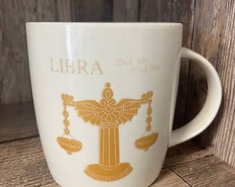 Pode incluir: Caneca de cerâmica branca com um símbolo de Libra do zodíaco dourado e as datas "23 de setembro - 22 de outubro".