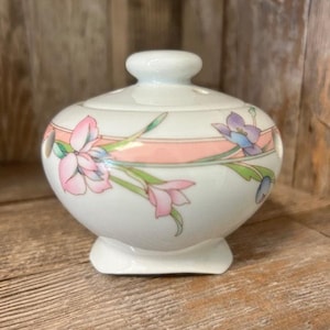 May include: A white ceramic jar with a lid, featuring a floral design with pink, blue, and green flowers. The jar has a pink and white stripe around the middle.