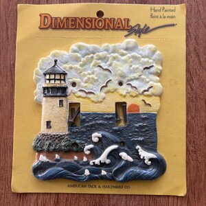 May include: A Dimensional Style double light switch plate featuring a hand-painted lighthouse scene with a sunrise, clouds, and crashing ocean waves. The packaging includes the text Dimensional Style, Hand Painted, and American Tack & Hardware Co.