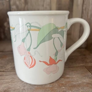 May include: White ceramic mug with a floral design featuring pink and orange flowers with green leaves. The flowers are arranged in a vine pattern around the mug.