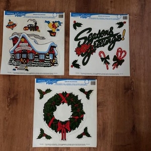 May include: Three vintage Christmas window clings. One features a Santa's workshop scene with a red roof and snow. Another says "Seasons Greetings!" with holly and candy canes. The third has a green wreath with a red bow.