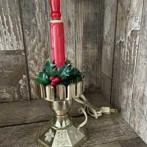 Puede incluir: Una vela eléctrica vintage de color dorado con una vela de plástico roja y acentos de acebo verde y bayas rojas. La vela está sobre una base decorativa con un cable y un enchufe.