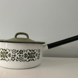 May include: White enamel saucepan with a black handle and a green floral design on the side. The lid is also white with a green rim.