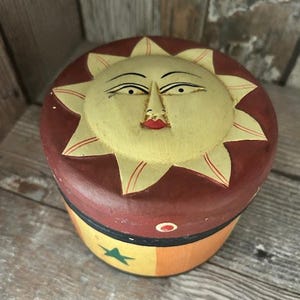 Könnte beinhalten: Eine runde, keramische Schmuckdose mit einem bemalten Design einer Sonne mit einem Gesicht. Die Sonne ist gelb mit einem roten Mund und grünen Augen. Die Dose ist rot mit einem gelben, orangen und schwarzen Streifen um den Boden. Ein grüner Stern ist an der Seite der Dose bemalt.