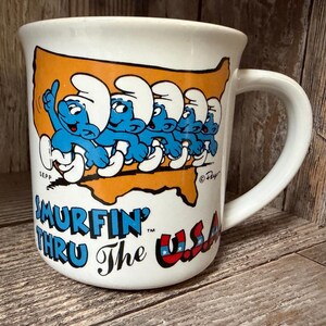 Puede incluir: Taza de cerámica blanca con un diseño de Pitufos azules marchando por un mapa de los Estados Unidos. La taza tiene el texto "Smurfin' Thru The USA" en azul y rojo, con los Pitufos en el centro.