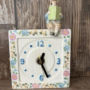 Puede incluir: Un reloj de pared cuadrado de cerámica con esfera blanca, números azules 3, 6, 9 y 12, y manecillas negras. El borde está decorado con un patrón floral de flores rosas, amarillas y azules. Una figura de conejo se asienta encima con un libro verde.
