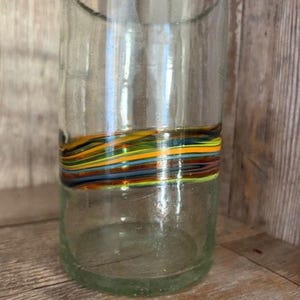 May include: A clear glass tumbler with a colorful, horizontal band of swirling stripes in shades of yellow, orange, blue, and green. The glass has a slightly textured appearance and sits on a wooden surface.