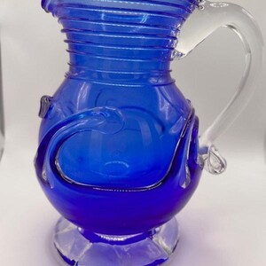 May include: A cobalt blue glass pitcher with a clear glass handle. The pitcher has a bulbous base and a ridged neck. Decorative swirls adorn the body. The pitcher sits on a clear, faceted base. A classic piece for serving beverages.