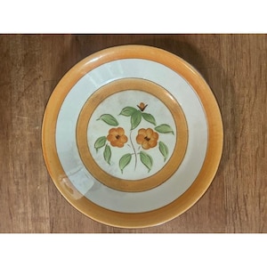 Puede incluir: Plato redondo de cerámica con diseño floral. El plato presenta un centro color crema con flores naranjas y hojas verdes. El borde está bordeado por una banda naranja claro y una banda blanca. El plato está sobre una superficie de madera.