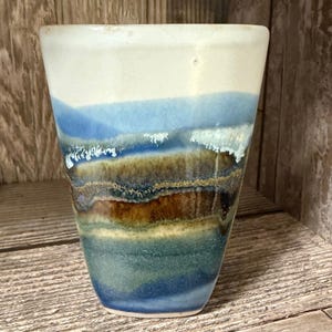 May include: A handmade ceramic tumbler with a unique, layered design. The top is white, transitioning to bands of blue, brown, and green. The textured surface gives the tumbler a rustic, artistic feel. This piece is ideal for enjoying beverages.