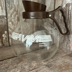 Puede incluir: Una cafetera de vidrio transparente con asa y tapa de plástico marrón. La cafetera tiene el texto "AVM COFFEE SERVICES" en blanco. La cafetera está sobre una superficie de madera.