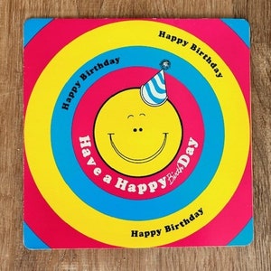 Könnte beinhalten: Eine bunte Geburtstagskarte mit einem Smiley, der einen Partyhut trägt. Die Karte hat konzentrische Kreise in Rot, Gelb, Blau und Pink mit dem Text "Happy Birthday" und "Have a Happy Birthday", der sich an den Rändern wiederholt.