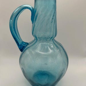 Puede incluir: Una jarra de vidrio azul con cuello retorcido y asa. La jarra tiene una superficie texturizada y está hecha de vidrio reciclado.
