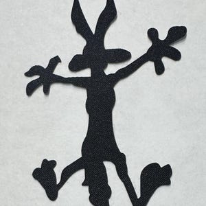 May include: A black fabric applique shaped like a cartoon coyote in a splayed, falling pose. The silhouette features long ears, outstretched arms with wide fingers, and legs bent at the knees, showcasing a textured, woven material surface.
