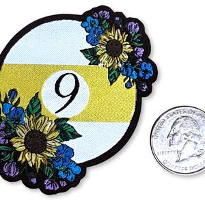 May include: A yellow and white embroidered patch with a floral design and the number 9 in the center. The patch is about the size of a US quarter.