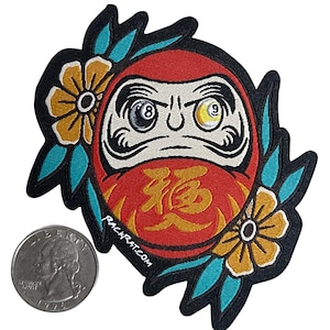 May include: A red and gold Daruma doll with a white face and black eyebrows. The doll's eyes are black and white billiard balls with the numbers 8 and 9. The doll is surrounded by yellow flowers and blue leaves. The text "福" is written in gold on the doll's belly. The text "rackrat.com" is written in white on the bottom of the doll.