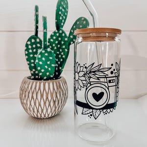 May include: A clear glass reusable water bottle with a bamboo lid and a black and white design of a camera with a heart in the lens. A glass straw is in the bottle. The bottle is next to a potted cactus.