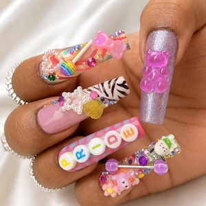Può includere: Unghie finte lunghe con varie decorazioni 3D, tra cui un orsetto gommoso rosa, un lecca-lecca arcobaleno, una stella di perle bianche e perline con lettere che compongono la parola PRIDE, su motivi colorati e finiture glitterate.