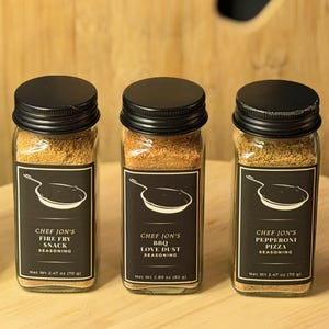 May include: Three glass spice jars with black lids and labels. The labels read "Chef Jon's Fire Fry Snack Seasoning", "BBQ Love Dust Seasoning", and "Pepperoni Pizza Seasoning". Each jar contains a different colored spice blend.