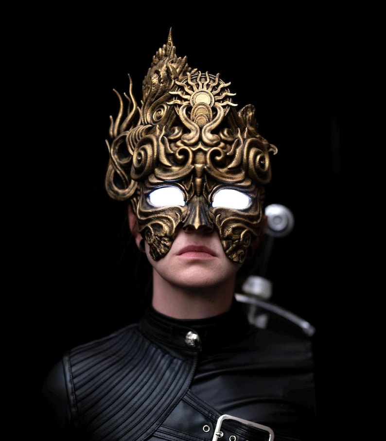 May include: A person wearing a gold, ornate mask with glowing white eyes. The mask has a detailed design with intricate patterns and a crown-like top.