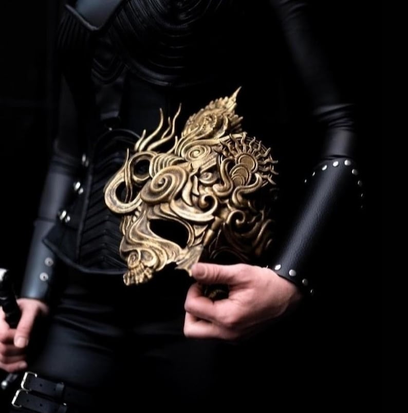 May include: A gold-colored mask with intricate details and a dragon-like design. The mask is held in the hand of a person wearing black clothing.