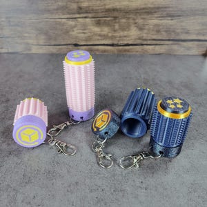 May include: A collection of 3D-printed keychains in various colors. The keychains feature cylindrical designs with textured surfaces and decorative tops. Colors include pink, purple, blue, and gold. Each keychain has a silver clasp.