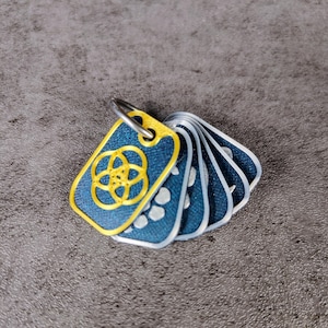 May include: A set of blue rectangular tags with a gold-colored design and a silver-colored ring. The tags have a denim-like texture and a gold border. The design on the first tag is a gold-colored floral pattern.