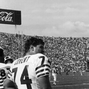 May include: Black and white photo of a football game. Two football players are wearing jerseys with numbers on them. The players are standing on a field with a large crowd of people in the background. A Coca-Cola sign is visible at the top of the image.