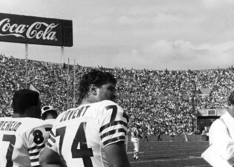 May include: Black and white photo of a football game. Two football players are wearing jerseys with numbers on them. The players are standing on a field with a large crowd of people in the background. A Coca-Cola sign is visible at the top of the image.