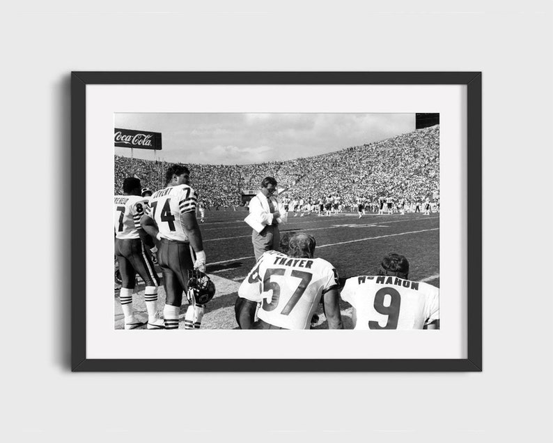 May include: Black and white photograph of a football team huddled together on the field. The players are wearing their uniforms and helmets. The crowd is visible in the background.