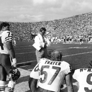 May include: Black and white photo of a football coach and players on the field. The coach is wearing a white shirt and tie, and the players are wearing jerseys with numbers. The photo was taken at a football game.